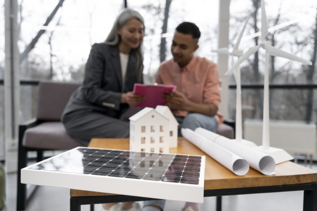 Energia solar. Maquete de casa, placa solar em miniatura e plantas enroladas em cima de mesa, em primeiro plano. Ao fundo, mulher e homem seguram e olham para prancheta rosa.