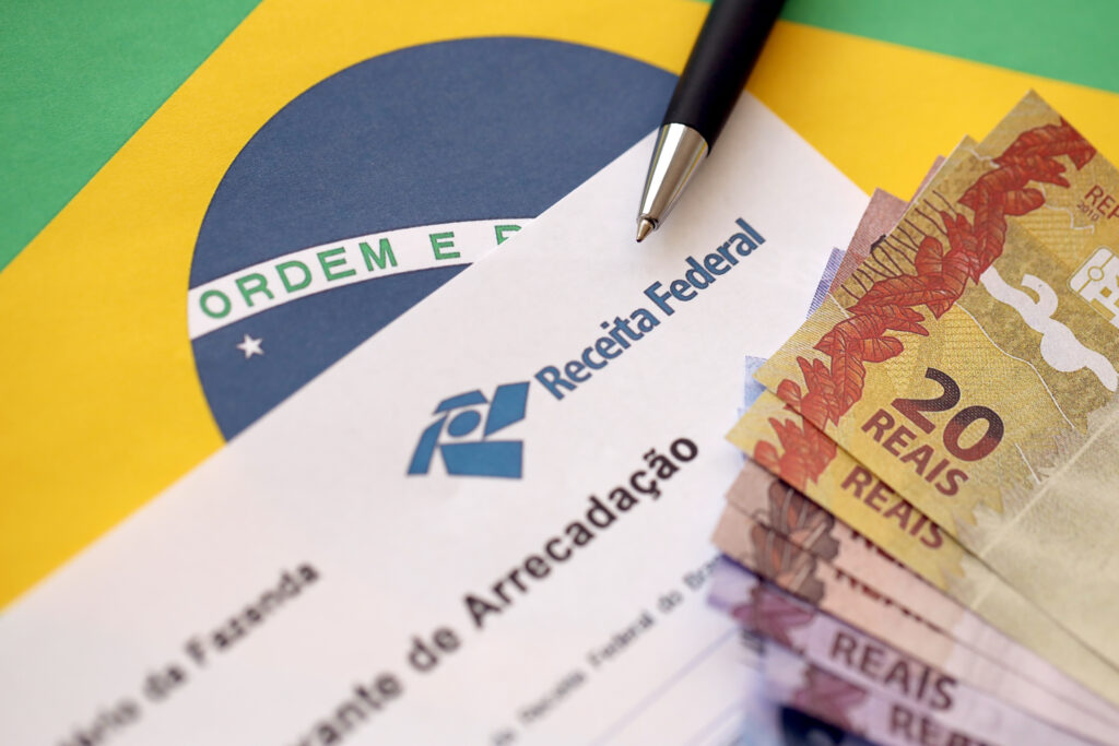 Imposto de Renda para autônomos, bandeira do Brasil ao fundo, notas variadas de dinheiro e papel da Receita Federal escrito "Comprovante de Arrecadação".