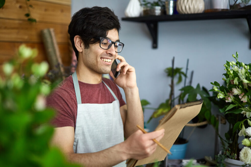 Imposto de renda para autônomos, interior de salão, homem de óculos usando avental fala ao celular e segura um grande bloco de notas mais um lápis.
