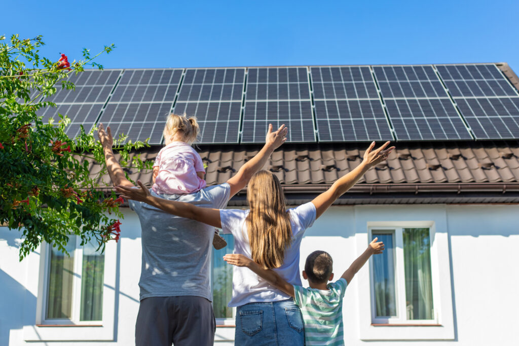 Soluções sustentáveis para casa própria. Família feliz, de pé e de costas, em frente a uma casa azul com muitas placas solares no telhado.
