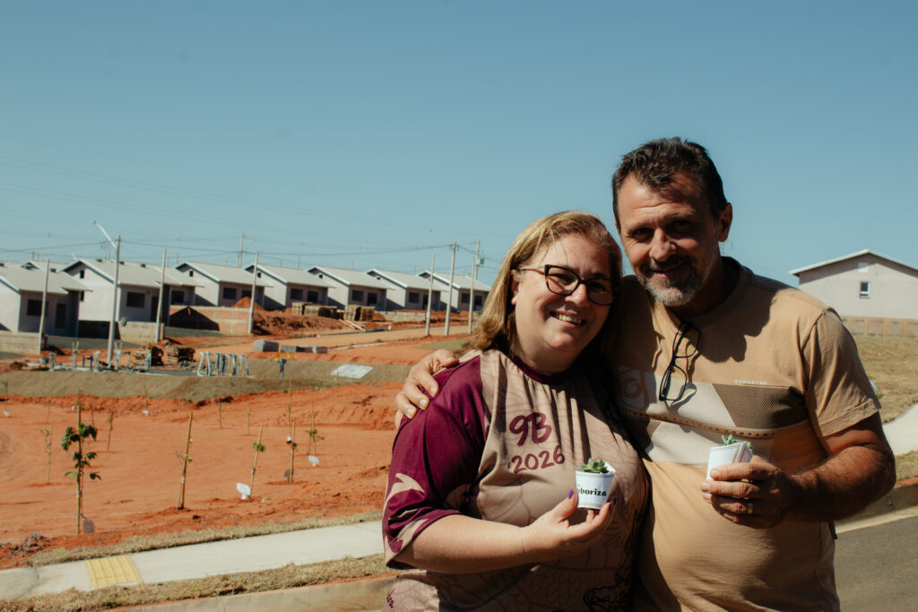 Imagem retangular de um casal, homem e mulher, posando para a foto abraçados e segurando um vaso pequeno de suculentas cada. Ao fundo, vê-se parte da obra do Residencial Monte Verde.