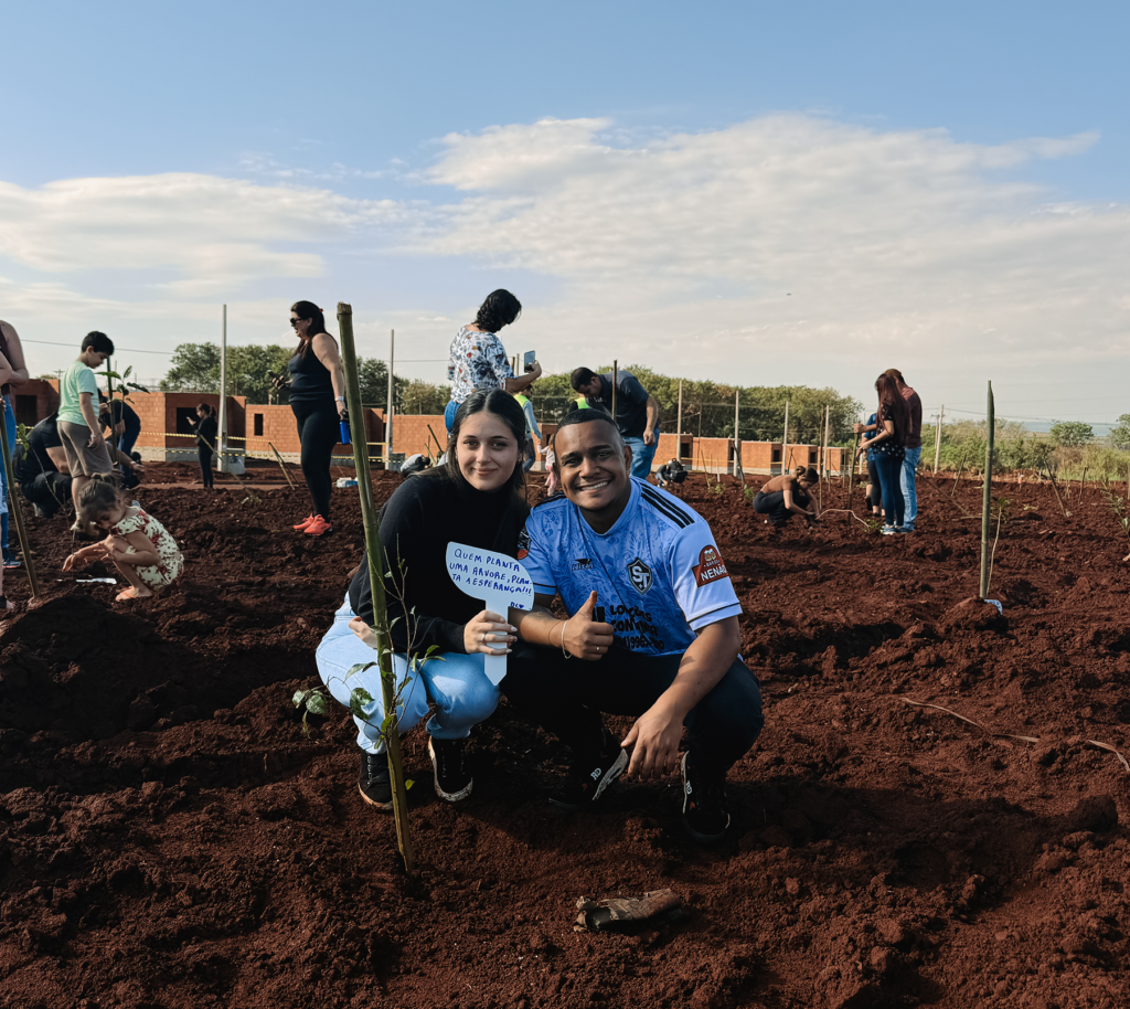 Foto de um casal, um homem negro e uma mulher branca, agachados ao lado da sua muda recém-plantada em um terreno amplo. Ambos estão sorrindo e ela segura uma plaquinha branca com a mão direita com os escritos "Quem planta uma árvore, planta a esperança!!".