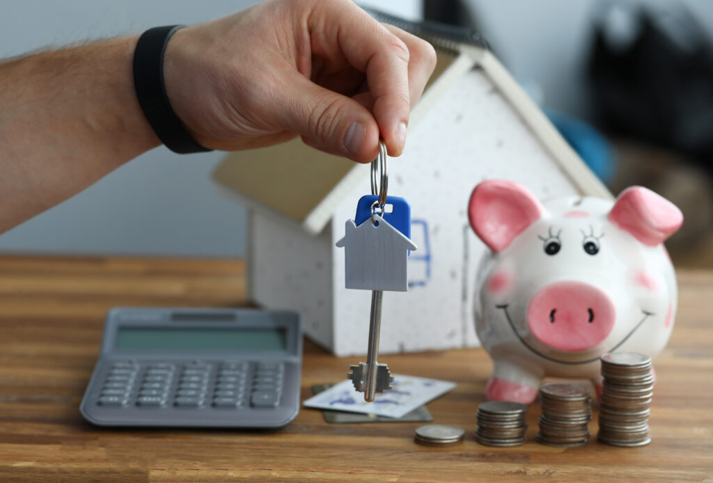 Economizar para comprar um imóvel, mão em primeiro plano, segurando chaves de casa; cofre de porquinho rosa e miniatura de casa ao fundo.