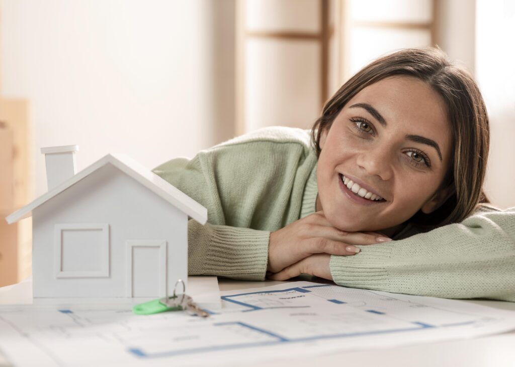 Economizar para comprar um imóvel. Miniatura de casa do tipo maquete de gesso, com uma mulher branca e sorridente, cabelos-longos, lisos e castanho-claro está bem próxima da maquete, apoiando-se na mesma superfície em que está a maquete.