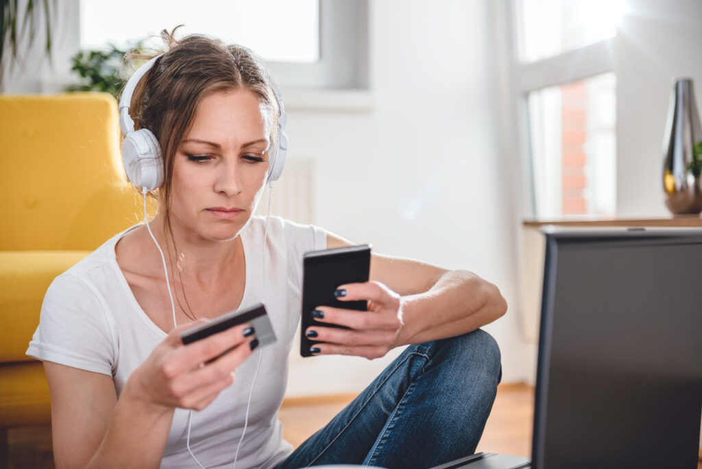 Economizar para comprar um imóvel. Mulher branca de calça jeans e camiseta branca, usando fones de ouvido do tipo headfone, sentada no chão, segurando cartão do branco em uma mão, celular na outra. 