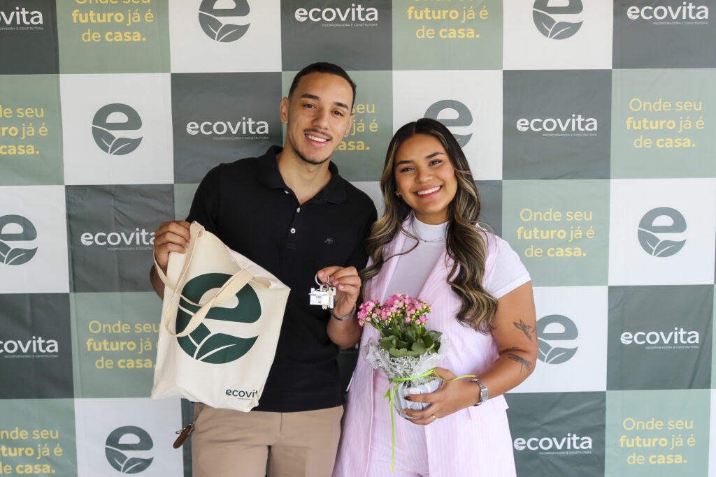 Foto de um um homem e uma mulher em frente ao backdrop da Ecovita. Ele é branco, veste uma camisa preta e uma calça bege e segura a ecobag da Ecovita na mão esquerda. Na mão direita, segura a chave. A mulher, também branca, está ao seu lado, com uma blusa branca e um blazer rosa claro, Ela segura, com a mão direita, um vaso de flor. Ambos sorriem para a foto.