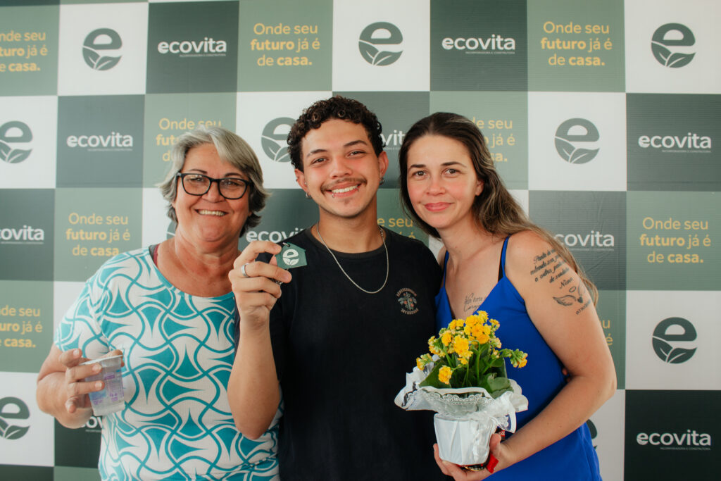 Foto retangular com 3 pessoas backdrop da Ecovita. A primeira é uma senhora mais velha, branca, de cabelos grisalhos curtos e de óculos de grau Ela veste uma blusa estampada branca e azul clara. Ao seu lado, está um rapaz mais novo, branco, de cabelos encaracolados escuros. Ele veste uma camiseta preta e uma corrente no pescoço. Com a mão direita, segura a chave da casa com o chaveiro da Ecovita. Ao seu lado, está uma mulher branca de cabelos longos e de luzes. Ela veste um vestido de alça azul e segura um vaso de flor amarela na mão esquerda. Os três estão sorrindo para a foto.
