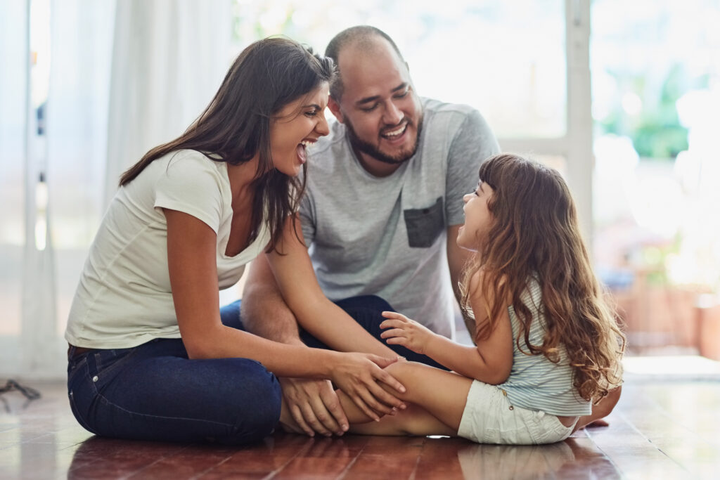 Momento ideal para comprar um imóvel. Foto de família de três integrantes, sentados no chão: mãe e pai distraem e brincam com filha criança.