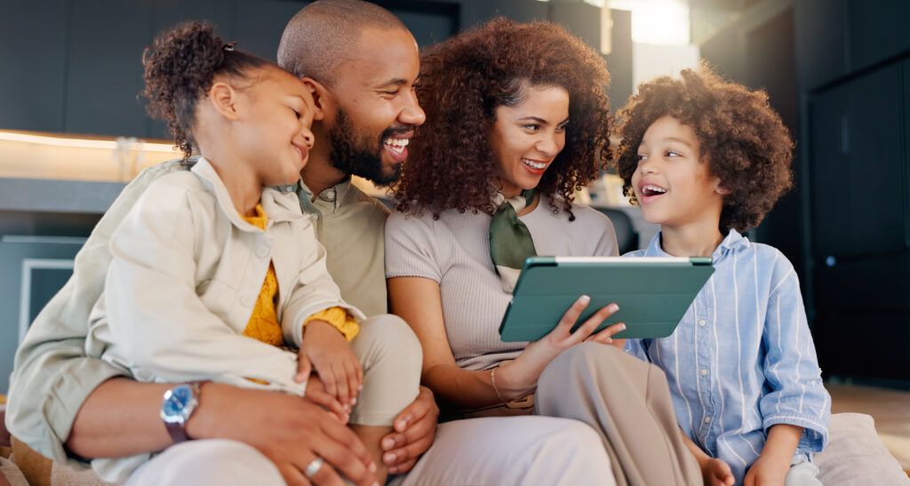 Planejamento financeiro para comprar um imóvel. Família brasileira, dentro de casa, pai, mãe, filho e filha sentados no sofá, todos olham tablet nas mãos da mulher, sentada no meio.
