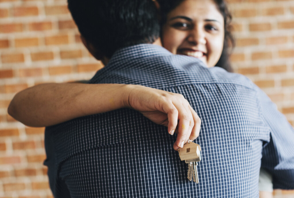 Vantagens de financiar um imóvel. Casal, homem e mulher, abraçados. Homem está de costas e mulher por trás dele, sorrindo e segurando com a mão direita a chave da casa própria.
