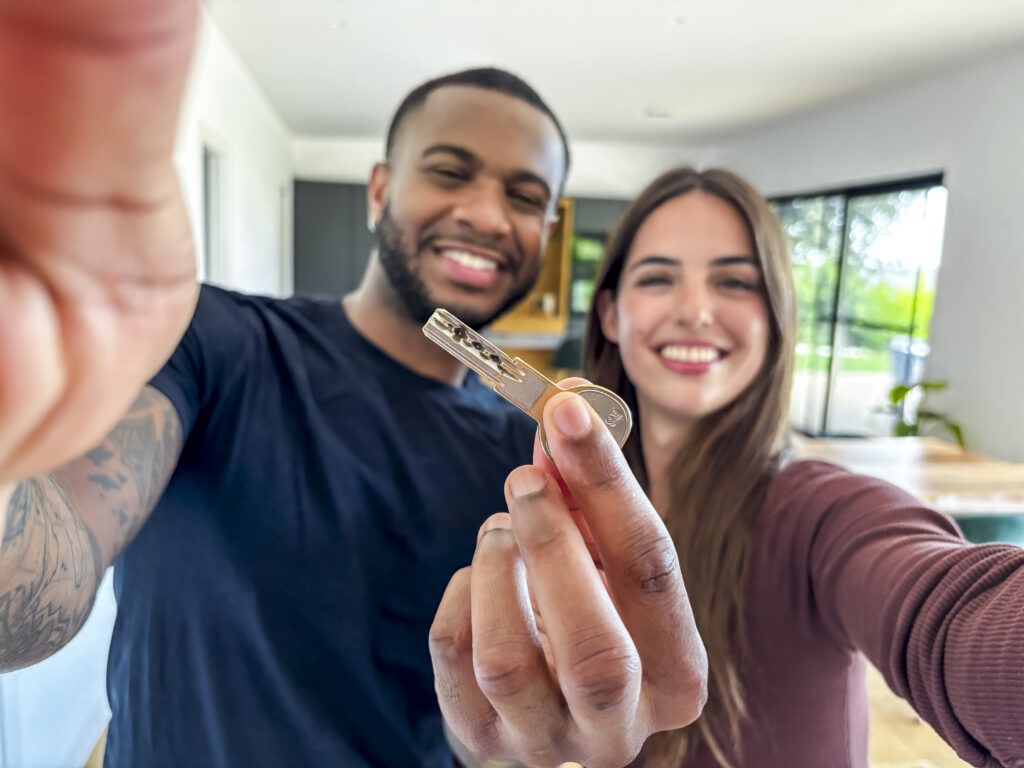 Vantagens de financiar um imóvel. Casal interracial, sorridente e feliz, segurando e olhando para câmera, em selfie. Homem, à esquerda, segura com a mão direita a chave da casa própria; mulher, à direita.