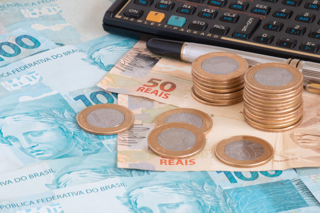 Vantagens de financiar um imóvel. Close de superfície plana repleta de notas de cinquenta e cem reais, junto com uma caneta, calculadora e pilhas de moedas de R$ 1,00.