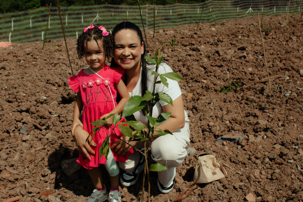 Foto de uma mulher e uma criança próximo a uma muda de árvore recém-plantada. A mulher veste uma roupa branca e a menina está com um vestido vermelho. A mulher está agachada, abraçando a menina pela cintura.