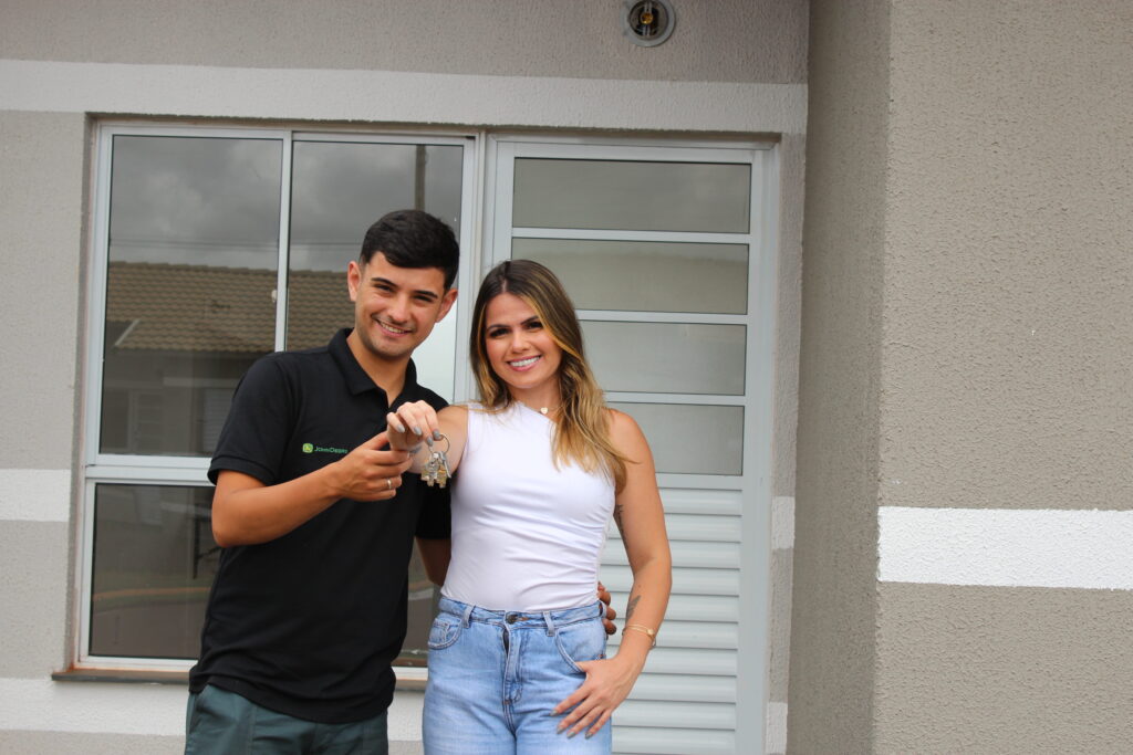 Foto de um casal em frente a casa da Ecovita. Ela é morena com luzes, tem olhos escuros e veste uma blusa branca de um ombro só e calça jeans. Ela está com a mão direita no bolso. Ele é moreno, de olhos castanhos e veste uma camiseta preta, com uma calça verde escuro. Ambos estão sorrindo para a foto e seguram juntos a chave da casa em frente do corpo.