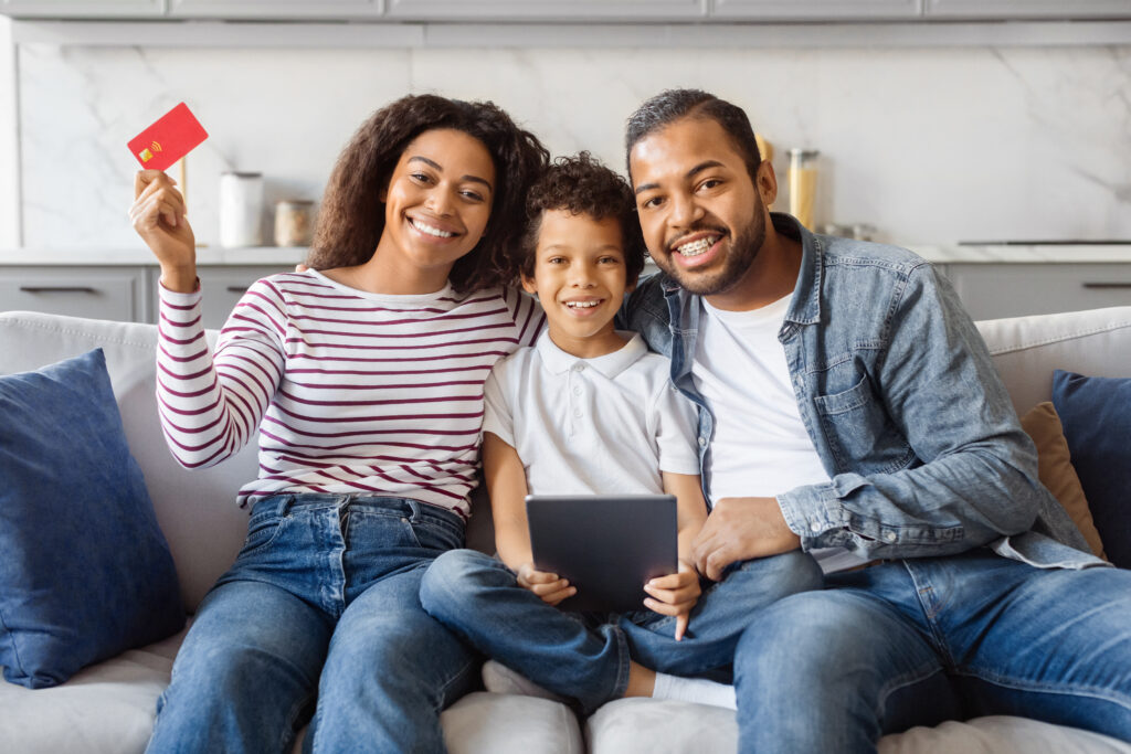 Análise de crédito imobiliário. Família de três, mãe, filho pequeno e pai, sentados no sofá de sala, felizes, sorriem para câmera. Mulher segura cartão na mão direita, estendida.