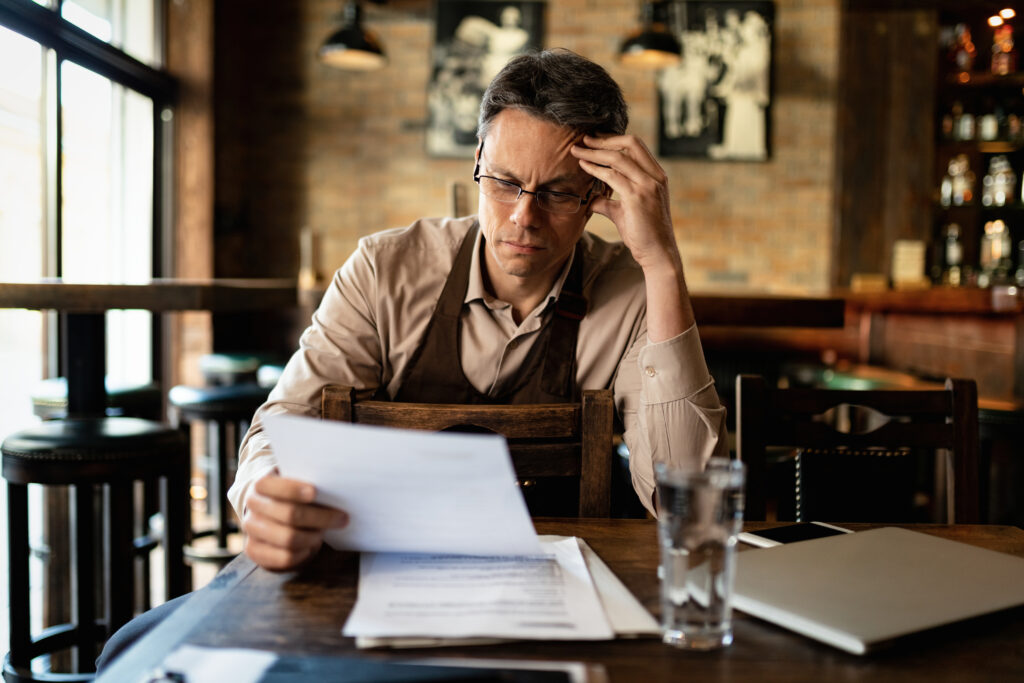 Análise de crédito imobiliário. Homem branco, empreendedor, vestindo um avental por cima da camisa, está sentado dentro de restaurante, olhando preocupado para papeis.