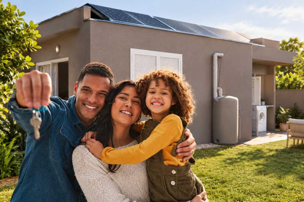 Valorização de imóvel. Foto de família de três, homem, mulher e filha pequena, todos felizes em frente à casa própria sustentável. Homem está segurando a chave da casa.